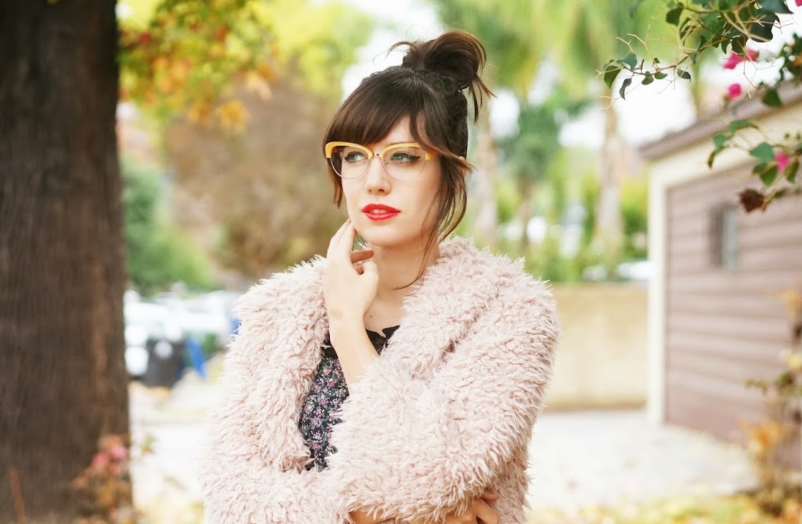 Woman wearing yellow-framed glasses, a fluffy pink coat, and red lipstick in an outdoor setting.