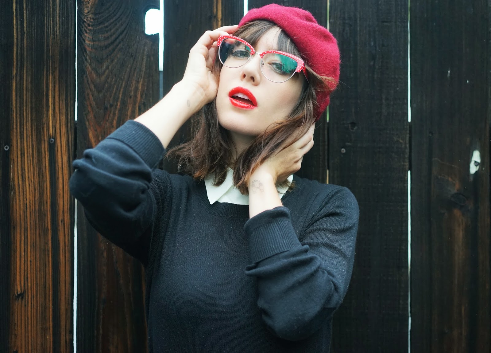 Woman wearing a red beret and red polka-dotted glasses with a black sweater and white collar.