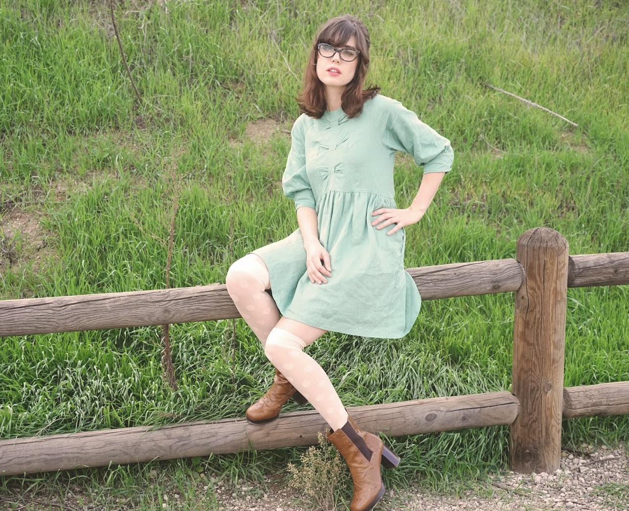 Woman in a green dress with polka dot stockings and brown boots sitting on a wooden fence.