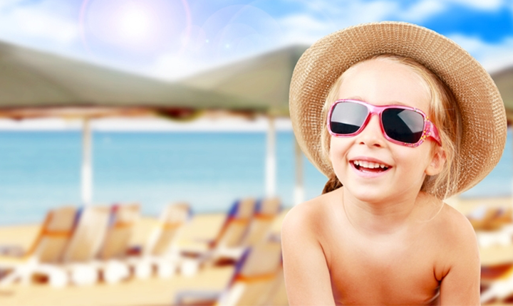 Smiling child wearing a sun hat and pink sunglasses on a bright, sunny beach.