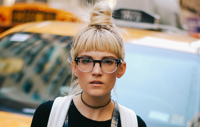 Blonde woman with a top knot wearing thick-rimmed glasses and a black choker necklace.