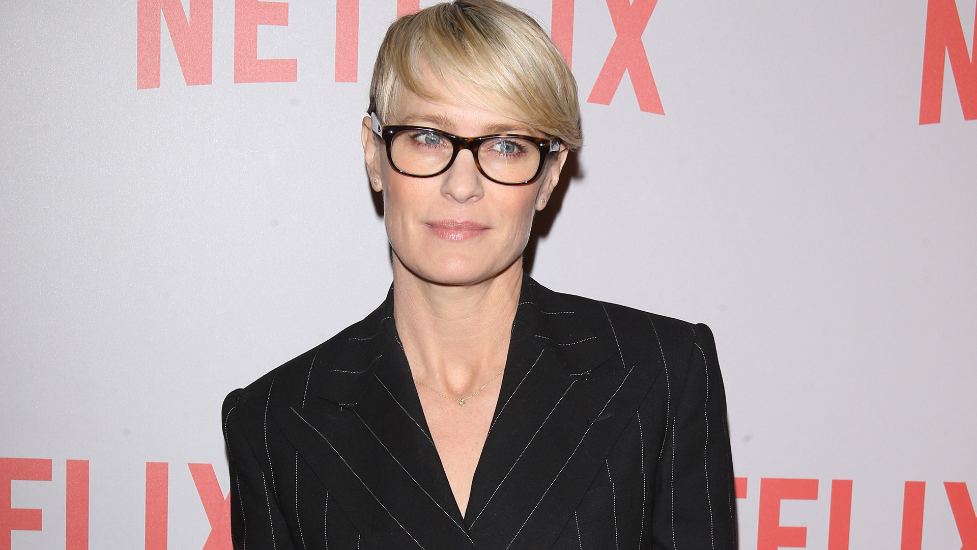 Person wearing black, pinstriped suit and black glasses. "NETFLIX" text is partially visible in the background.