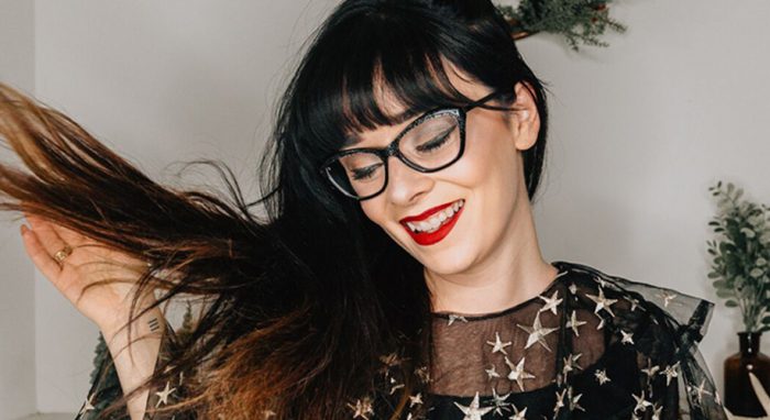 Woman wearing star-patterned sheer black top, large glasses, and red lipstick smiles while flipping hair.