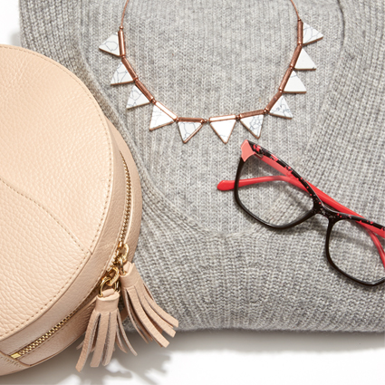 Beige round handbag, red and black eyeglasses, triangle necklace, and gray sweater in a flat lay arrangement.