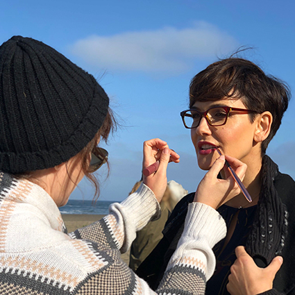 Person applying lipstick to another individual wearing glasses by the beach.