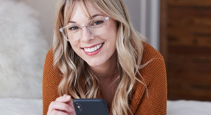 Woman wearing clear eyeglasses and an orange sweater, using a smartphone with a smile.