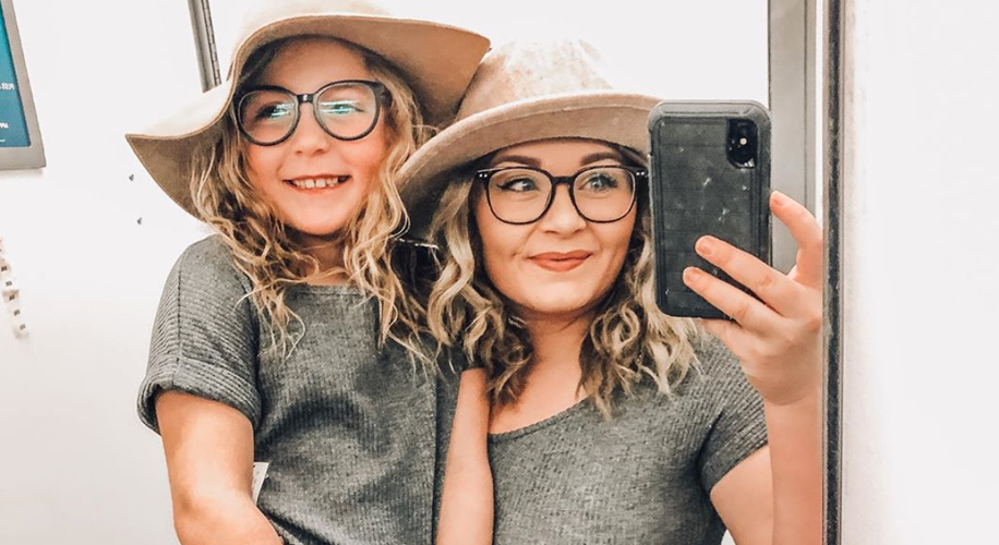 Woman and child wearing beige hats and grey outfits, smiling and taking a selfie in a mirror.