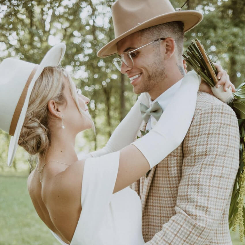 Couple in wedding attire smiling and embracing; bride holding a bouquet, both wearing hats.