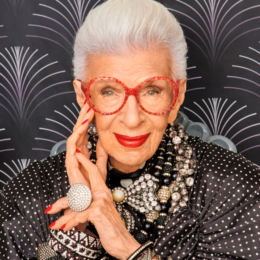 Woman with red glasses, wearing a beaded black outfit and accessories, smiles with hand on face.