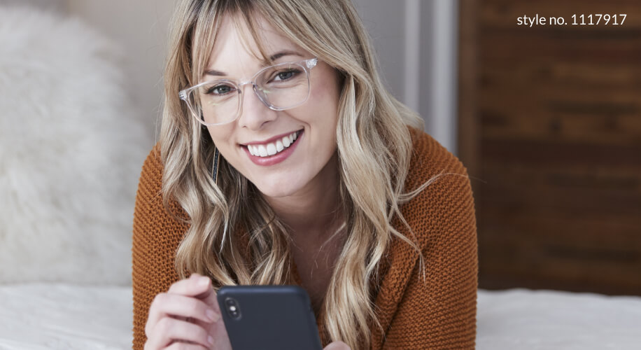 Woman wearing clear eyeglasses, style no. 1117917, smiling while holding a smartphone.