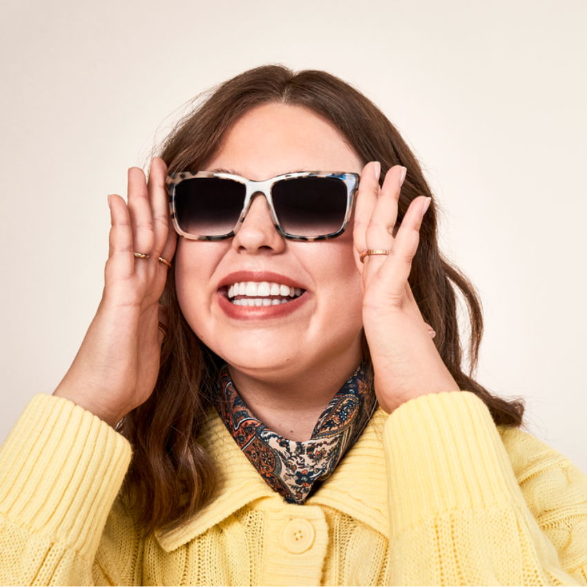 Woman smiling, wearing oversized square sunglasses with a tortoiseshell pattern and a yellow sweater.