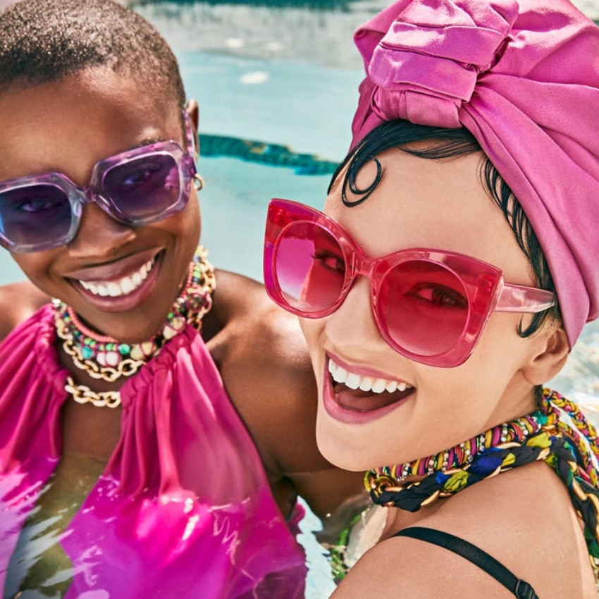 Women wearing large, colorful sunglasses with bright pink and purple frames, accessorized with vibrant clothing and hats.