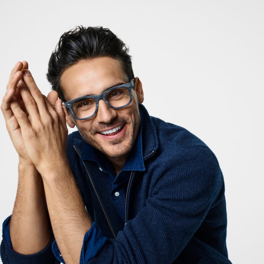 Man wearing blue glasses and a blue sweater, smiling with hands clasped.