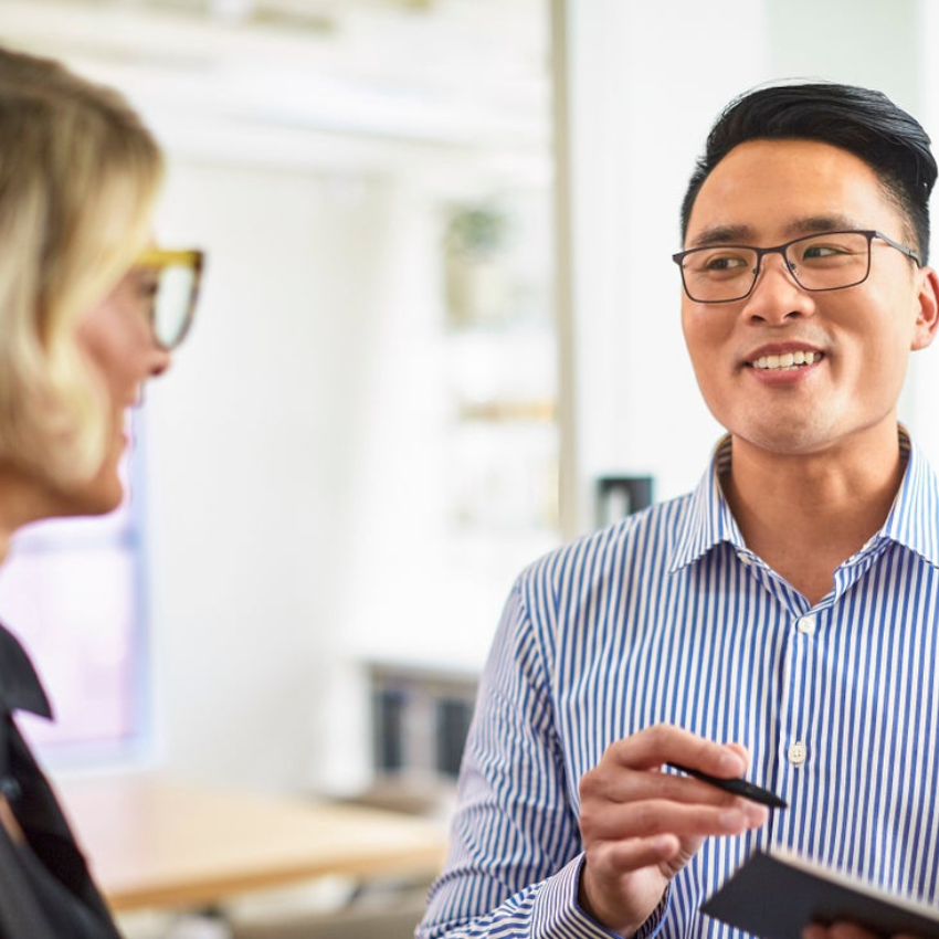 Man in striped shirt holding a notebook and pen, conversing with a woman wearing glasses.