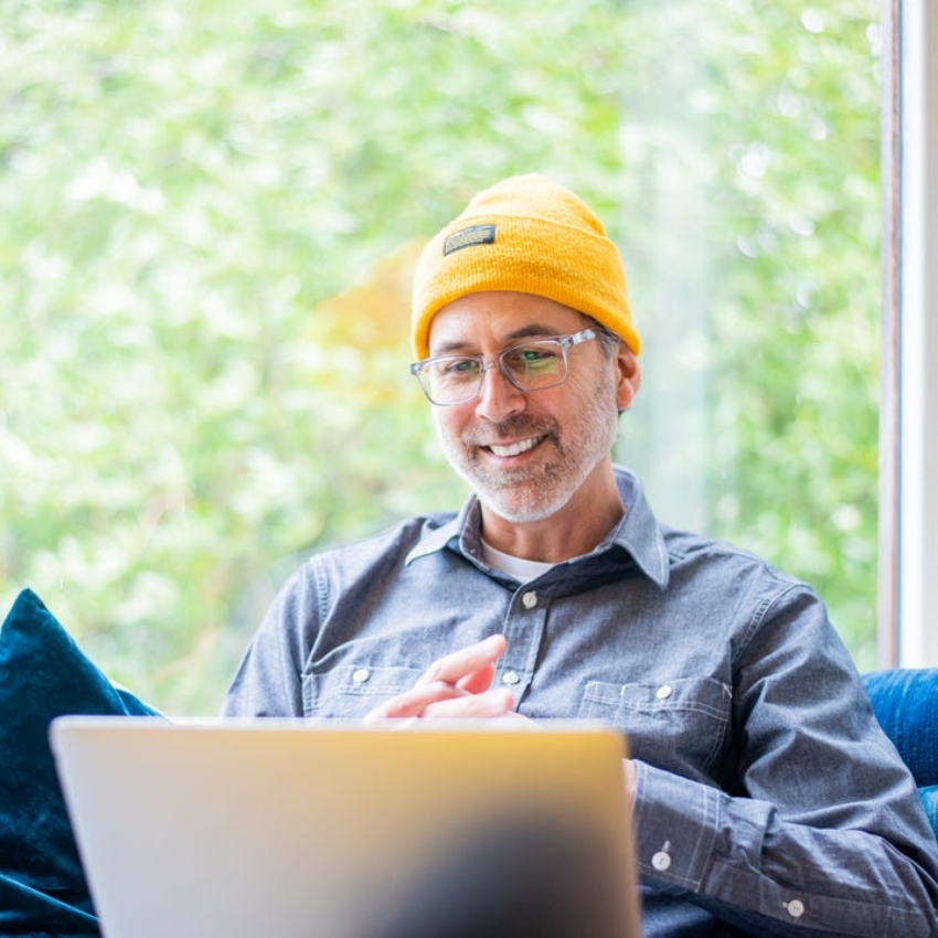 Smiling man wearing a yellow beanie and glasses, working on a laptop.