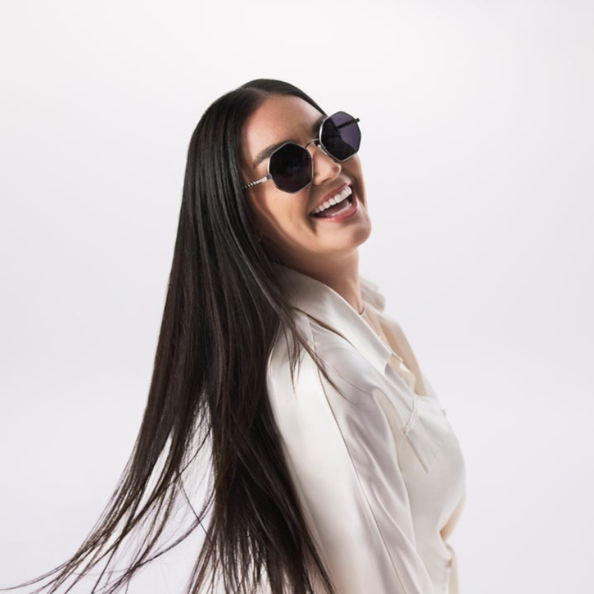 Woman wearing octagonal black sunglasses and a white blouse, smiling with long dark hair flowing down.