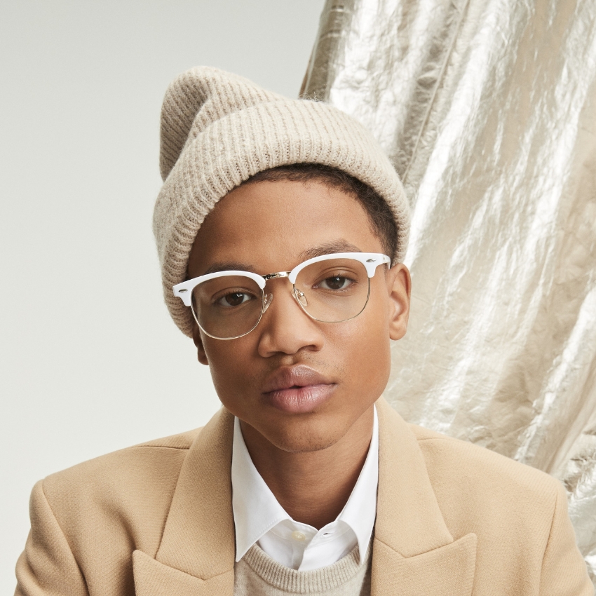 Person wearing beige knitted beanie, trendy white-rimmed glasses, beige coat, white shirt, and beige sweater.