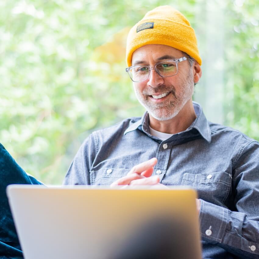 Person wearing a yellow beanie, glasses, and a gray button-up shirt, smiling while using a laptop.