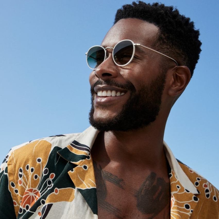 Man wearing round sunglasses with silver frames and a yellow floral-patterned shirt, smiling outdoors.