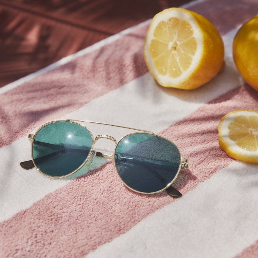 Gold-rimmed aviator sunglasses with teal lenses on a pink and white striped towel, lemons nearby.