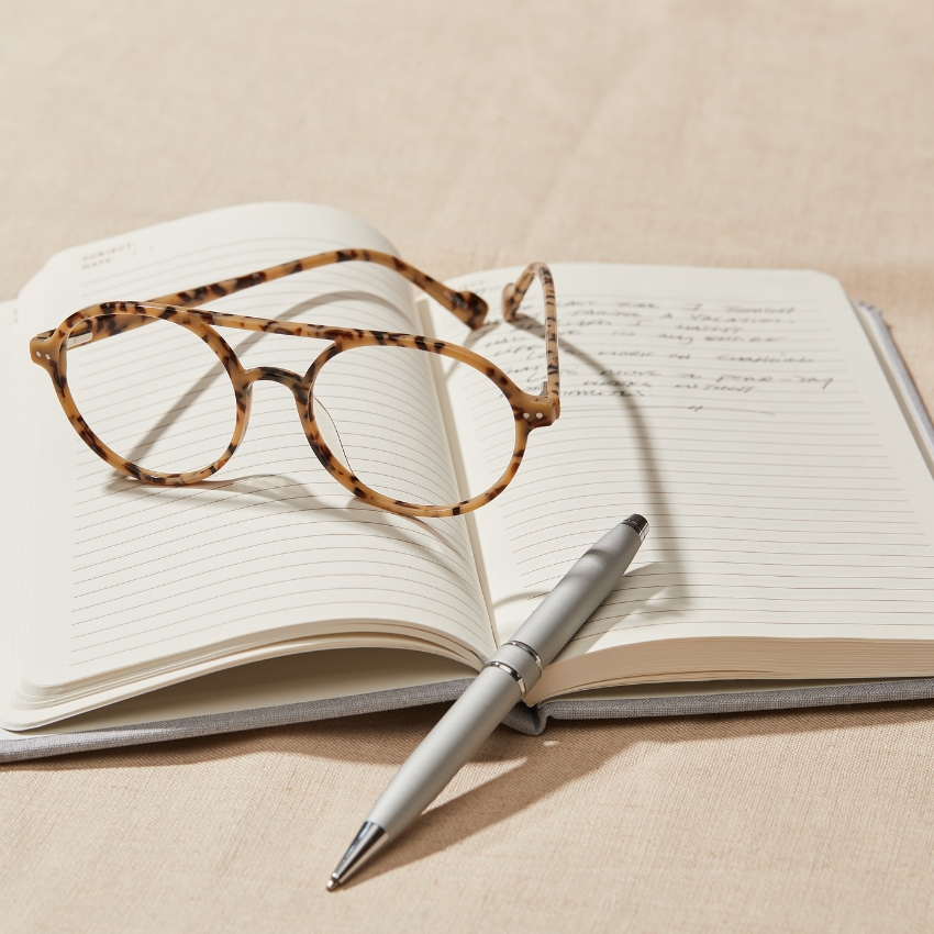 Tortoiseshell round glasses resting on an open notebook with handwritten notes, next to a silver pen.