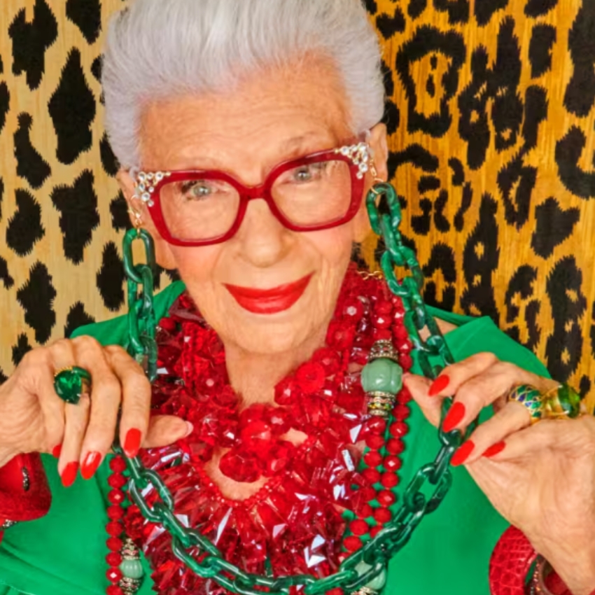 Woman wearing red glasses, red lipstick, large green and red bead necklaces, matching rings, and green top.