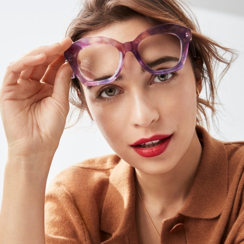 Woman wearing a brown jacket, holding purple translucent eyeglasses with one hand.