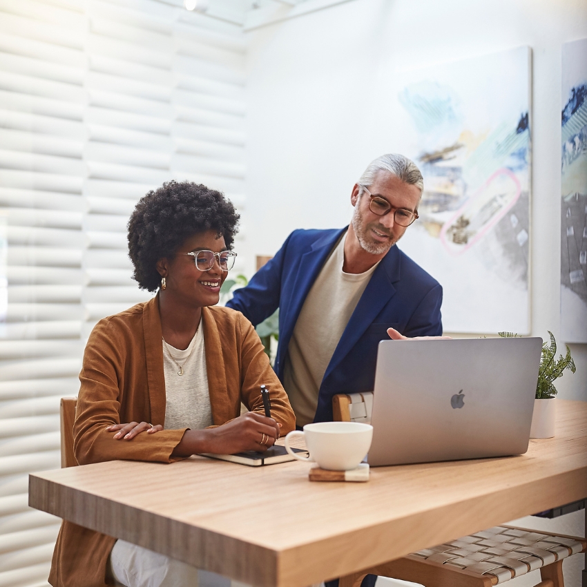 Two people discussing work on a grey open laptop with a coffee cup and notebook on the wooden table.