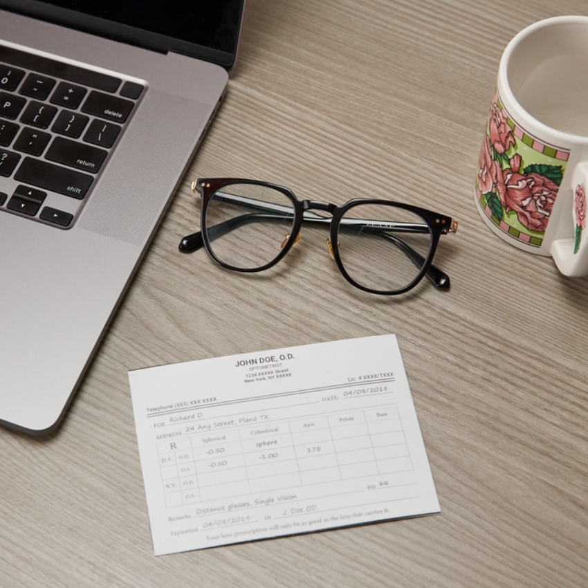 Eyeglasses placed on a wooden surface next to a laptop, prescription form, and a floral mug. Prescription visible is for Richard D., dated 04/08/2018, with Sphere, Cyl, and Axis details. Doctor’s name is John Doe, O.D.