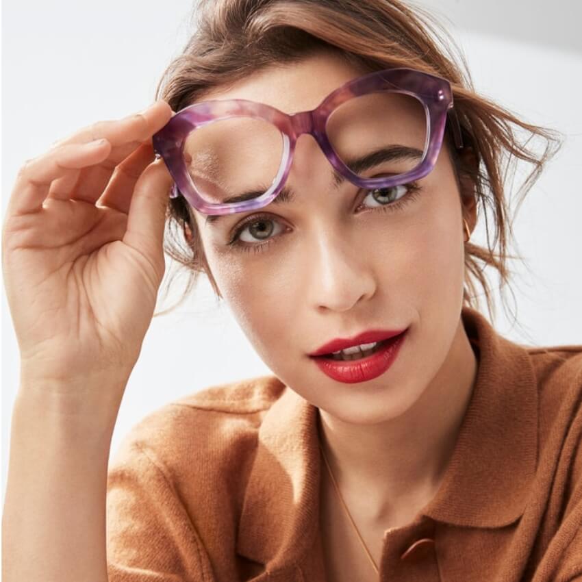 Woman wearing large, purple-framed eyeglasses, slightly lifted with one hand.