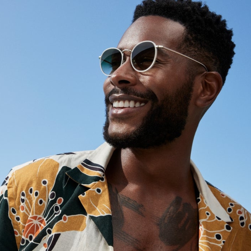 Man wearing round sunglasses and a floral print shirt smiles under a clear blue sky.