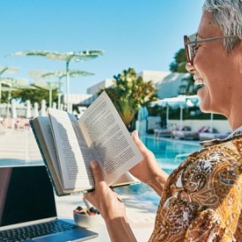 Person holding an open book near a poolside, with a laptop and bowl of fruit in the background.