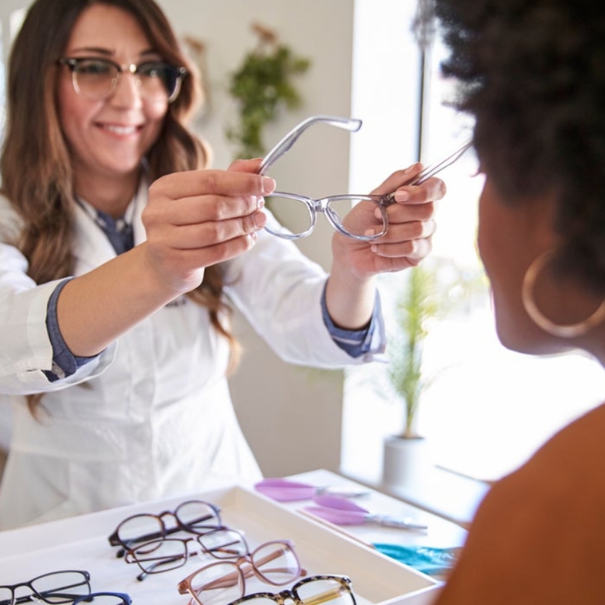 Optician showing clear eyeglass frames to a customer; various eyeglasses displayed on a tray.