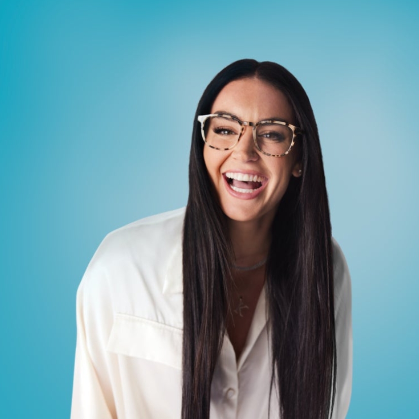 Woman wearing light tortoiseshell eyeglasses and an ivory shirt, laughing against a light blue background.