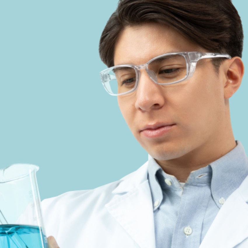 Person in a white lab coat wearing safety glasses, examining a beaker with blue liquid.