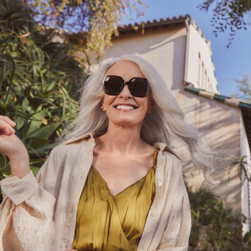 Woman wearing oversized black sunglasses and a yellow dress, smiling brightly outdoors.