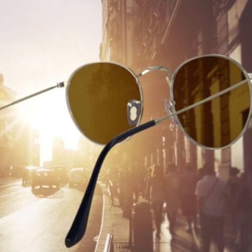 Round sunglasses with thin metal frames and brown-tinted lenses floating in front of a sunlit street background.