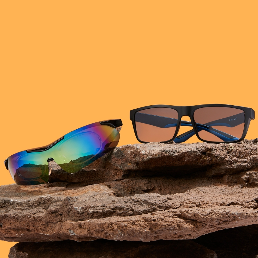 Sunglasses on rocks. One pair has rainbow mirrored lenses, the other has dark rectangular lenses.