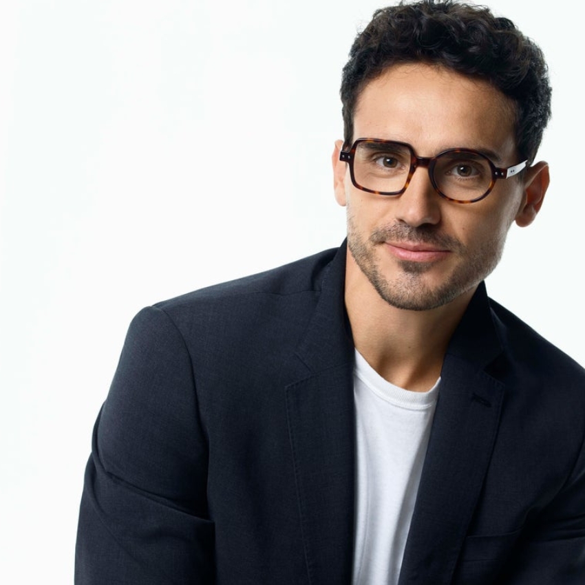 Man wearing tortoiseshell rectangular glasses, white t-shirt, and a dark blazer.