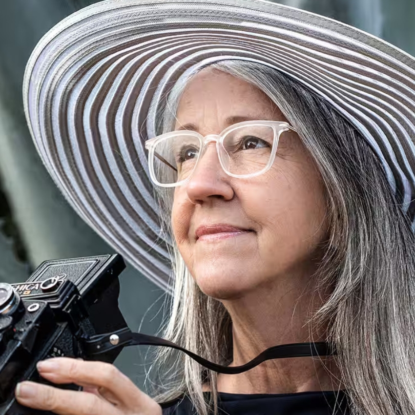 Woman wearing white-rimmed glasses and a wide-brimmed striped hat, holding a Yashica camera.