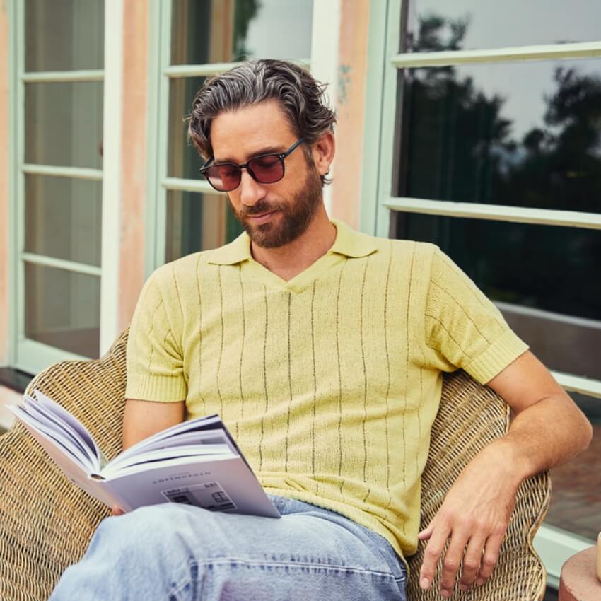 Man wearing a light yellow knit polo shirt and blue jeans, holding and reading a book while seated.