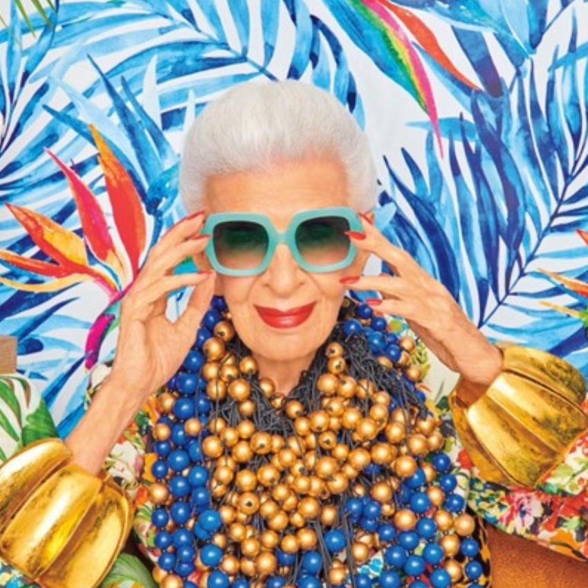 Woman wearing oversized turquoise sunglasses, large gold bracelet cuffs, and multiple blue and gold bead necklaces.