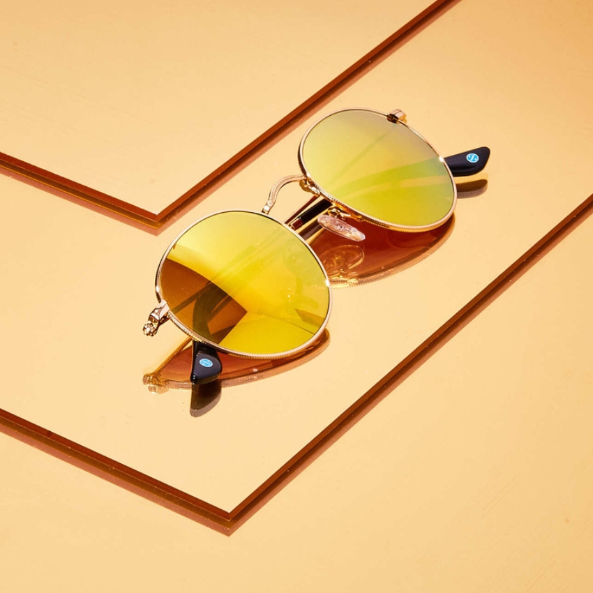 Sunglasses with round yellow lenses and gold frames on a reflective yellow surface.