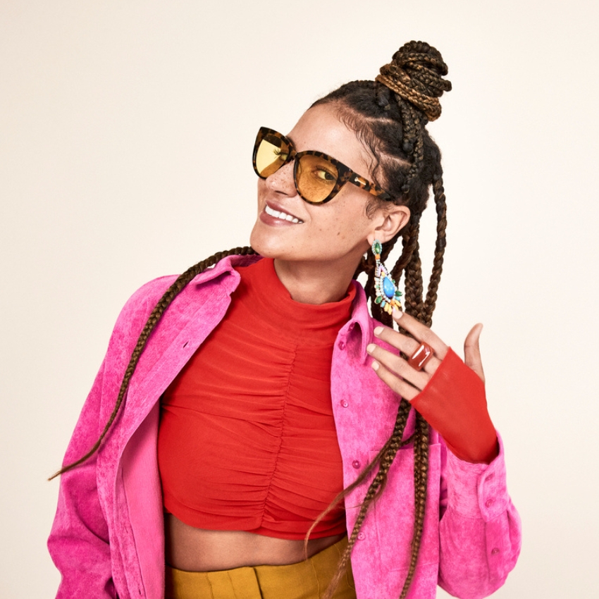 Woman wearing tortoiseshell cat-eye sunglasses, a red ruched top, pink jacket, and colorful earrings.