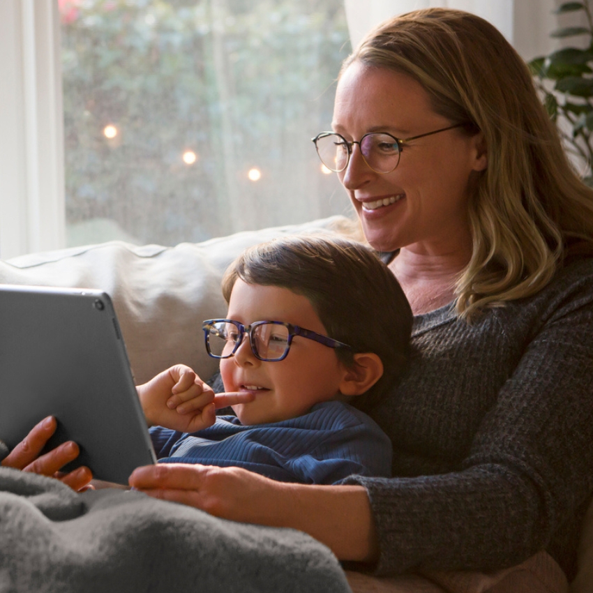 Mother and child wearing glasses, smiling while looking at a tablet together.