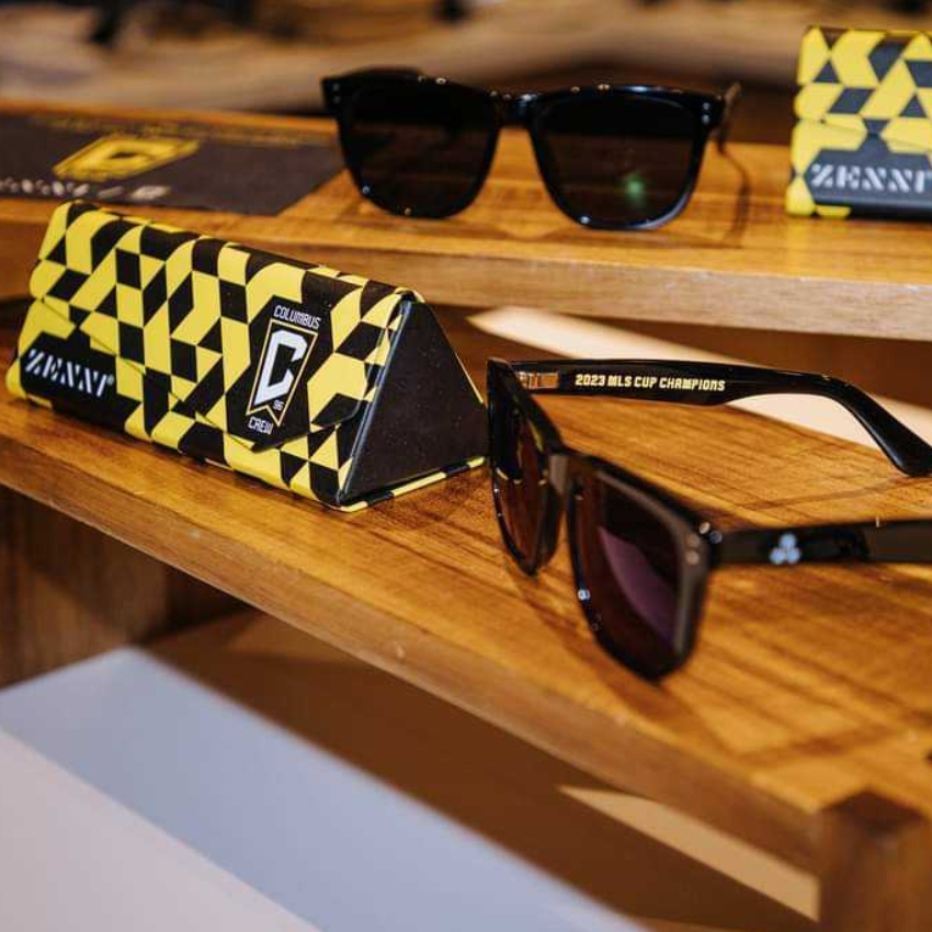 Black sunglasses with "2023 MLS CUP CHAMPIONS" on the arm. Columbus Crew logo on yellow checkered case.