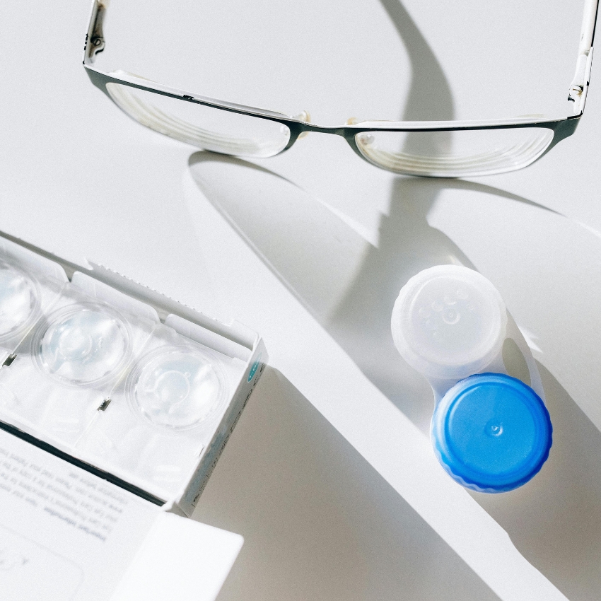 Glasses, a contact lens case, and a box of contact lenses on a white surface.