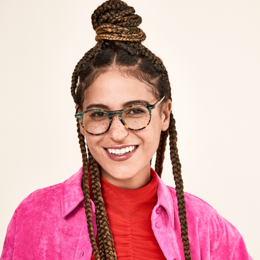 Person wearing tortoiseshell glasses with green accents, a pink jacket, and a red top, smiling.