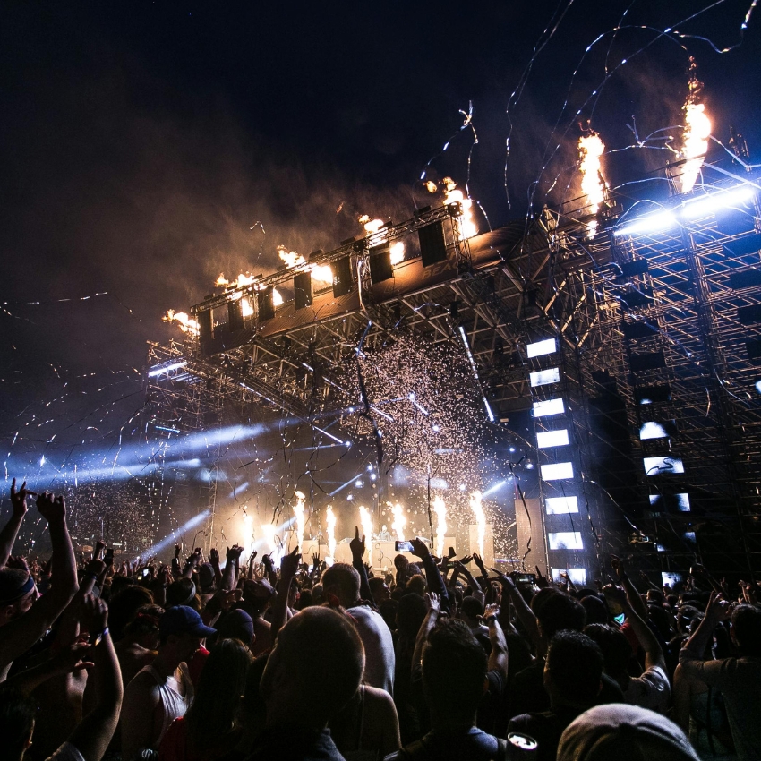 Concert stage with pyrotechnics, confetti, and lights, crowd with raised hands enjoying the performance.