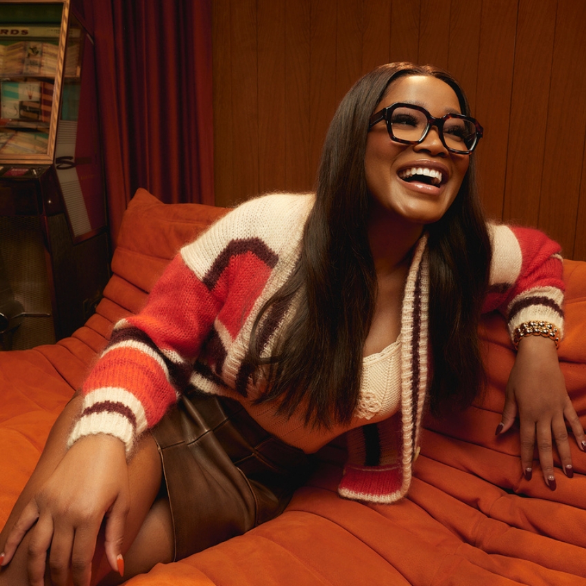 Person laughing, wearing geometric multicolored cardigan, white top, glasses, and brown leather skirt.